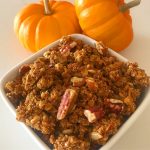 A white bowl filled with granola next to two pumpkins.
